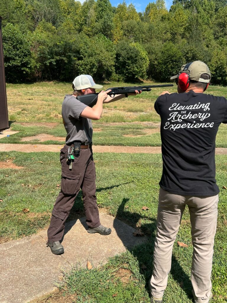 A person instructs another on shooting a gun in a field.