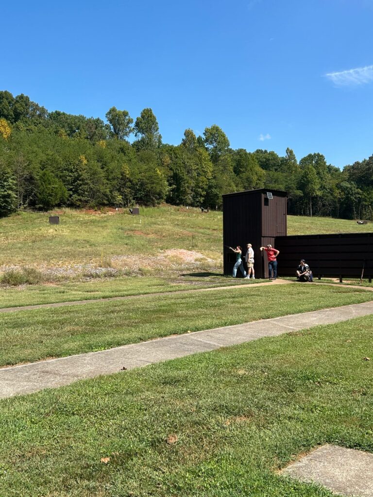 Two people stand and watch a person shoot a gun into a field.