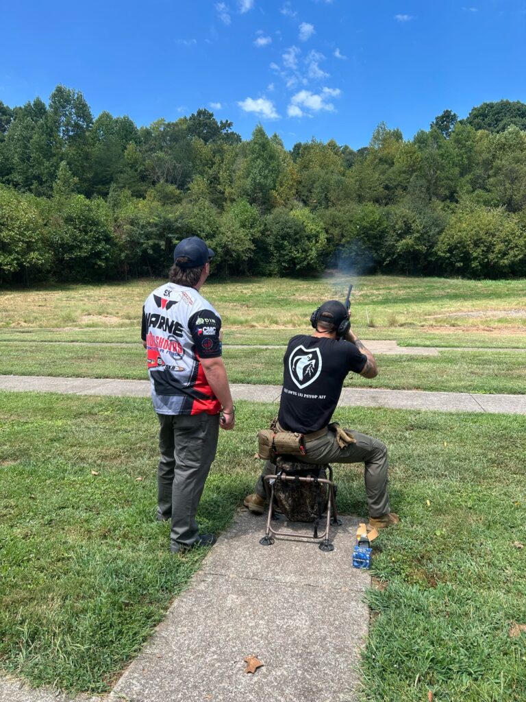 A person stands and watches another shooting a gun into a field.