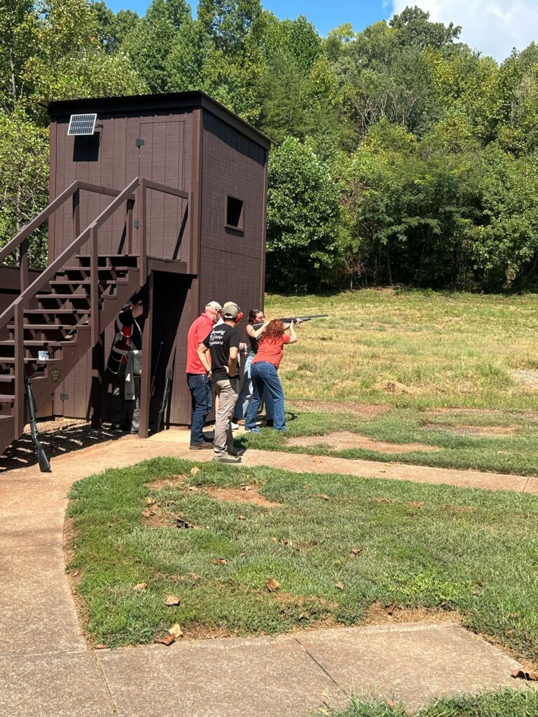 Two people watch a person shoot into a field with a building behind them.