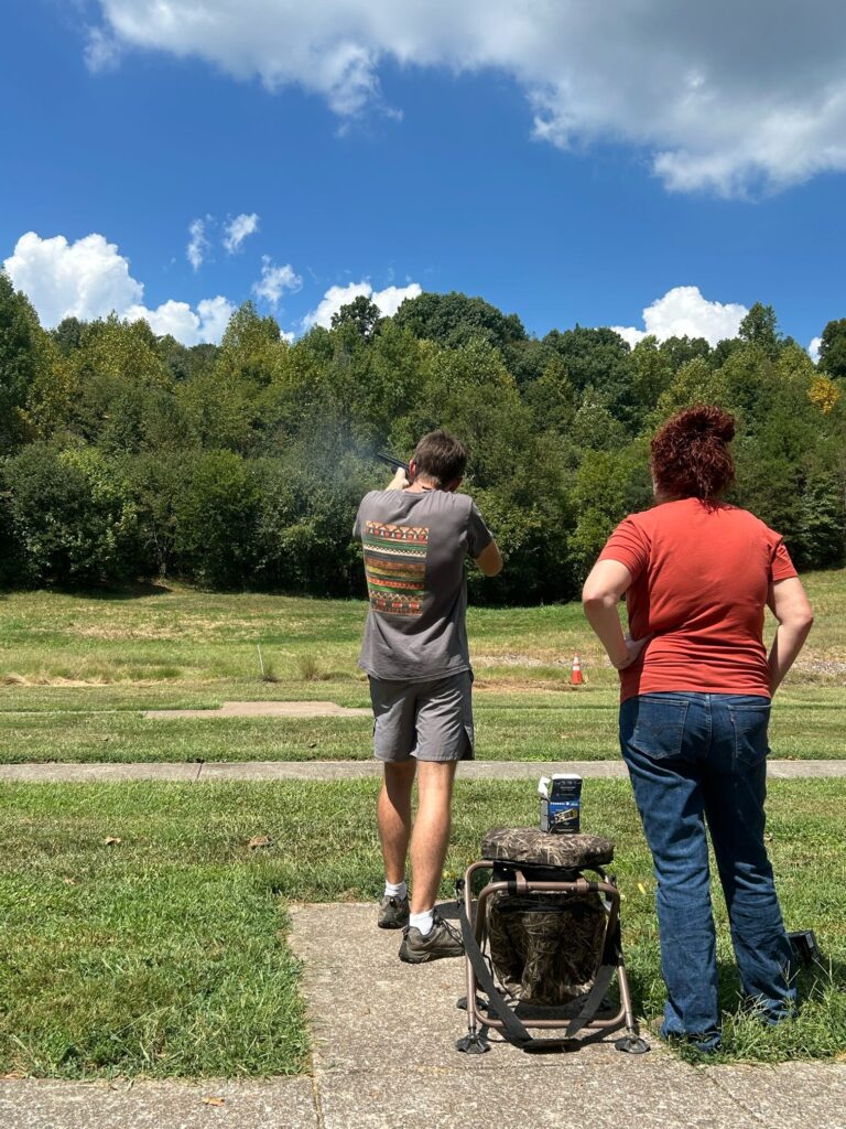 One person watches another shooting a gun into a field.