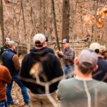 Twelve people listen to a person talk in the woods during the fall.