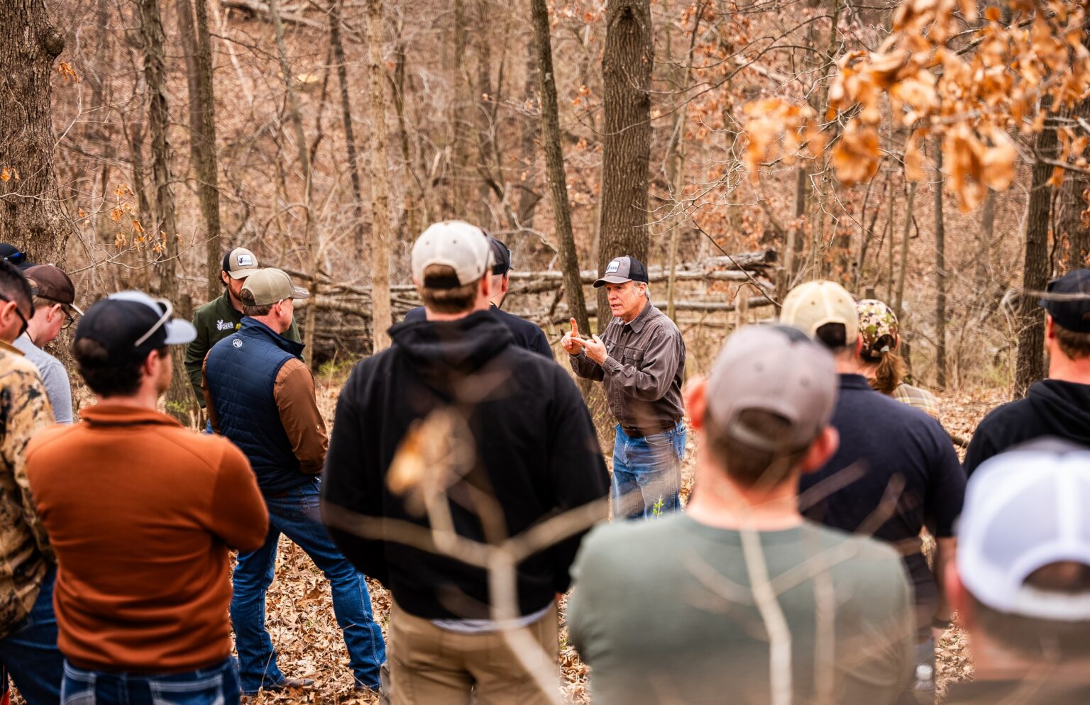 Twelve people listen to a person talk in the woods during the fall.