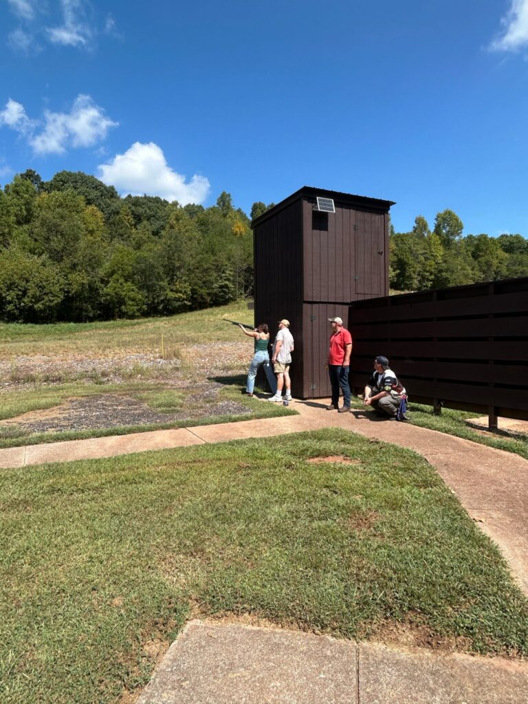 Three people watch a fourth shoot a gun in a field.