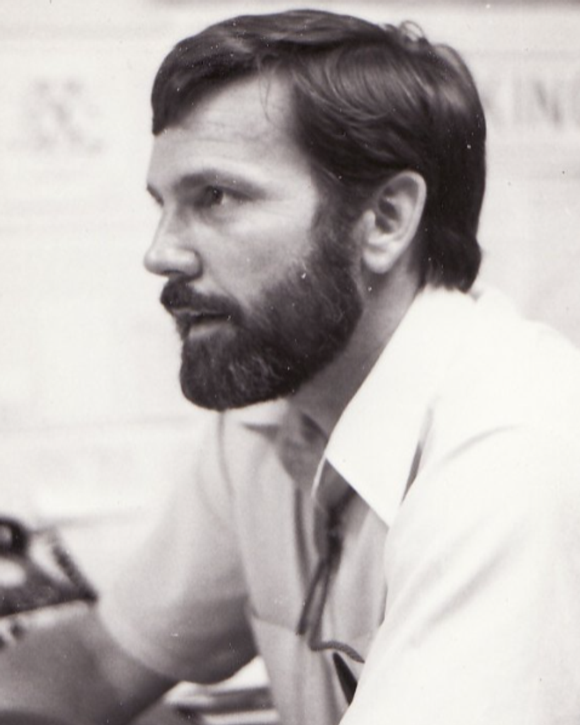 Black and white photo of bearded person wearing collared shirt.