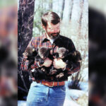 Person wearing flannel shirt holds four bear cubs in the woods.