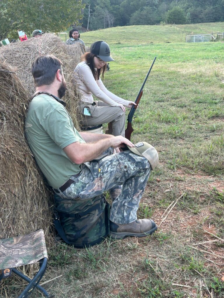 One person holds a gun next to another while sitting by a hay bale.