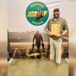 Man stands in front of banner for Tennessee Volunteer Educator Program holding wooden plaque.