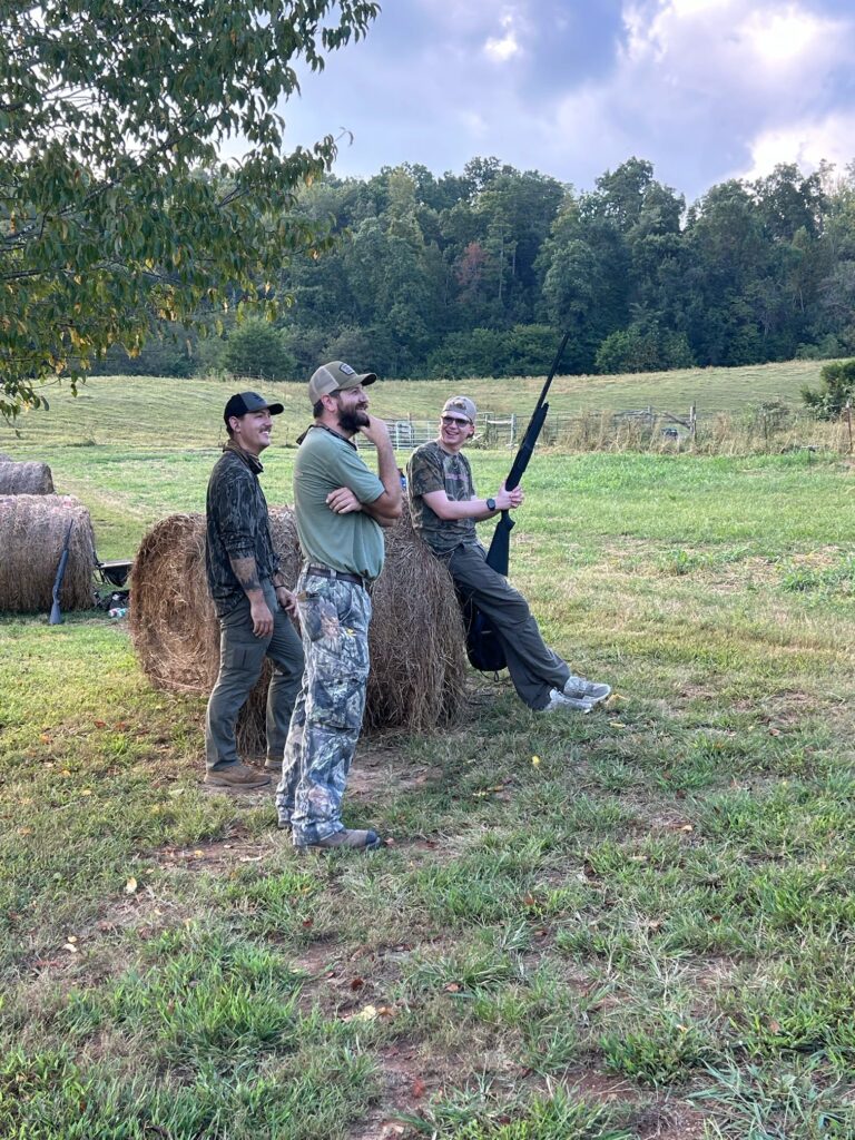 One person holds a gun and leans against a hay bale by two others.