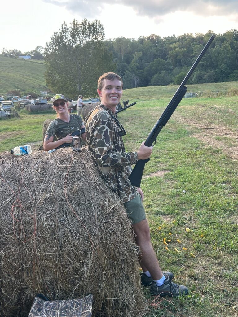 Two hunters stand beside a hay bale in a field.